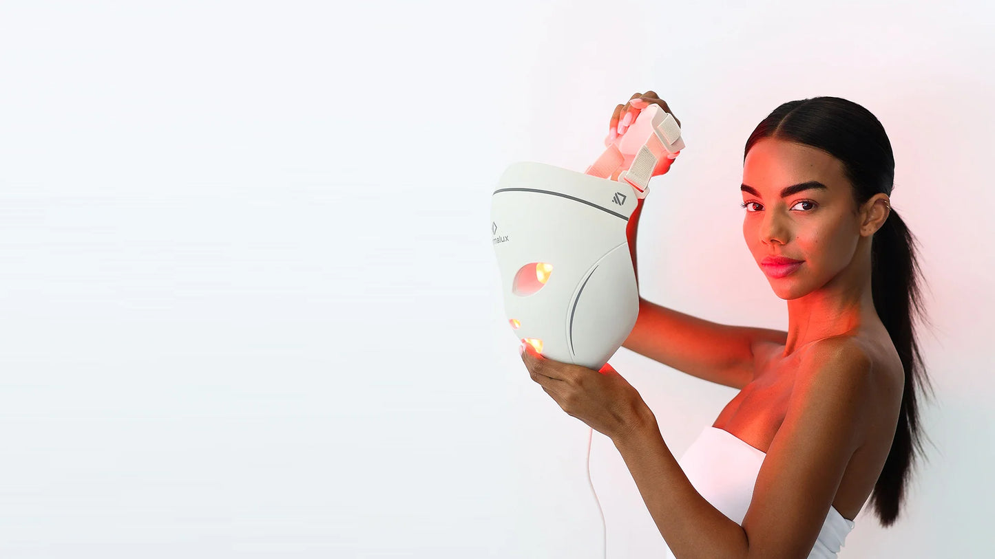 A woman holding the lit Dermalux LED Face Mask beside her face, red light falling across her shoulder and neck