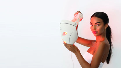 A woman holding the lit Dermalux LED Face Mask beside her face, red light falling across her shoulder and neck