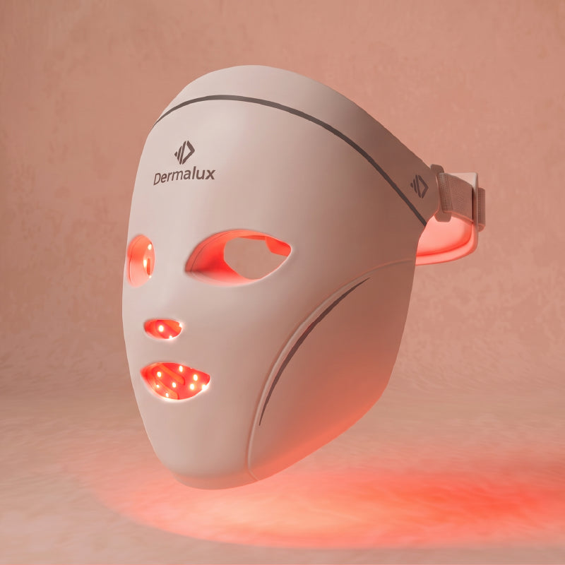 The Dermalux LED Face Mask at a three-quarter angle, red light glowing through the eye, nose and mouth openings, against a soft blush background