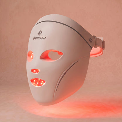 The Dermalux LED Face Mask at a three-quarter angle, red light glowing through the eye, nose and mouth openings, against a soft blush background