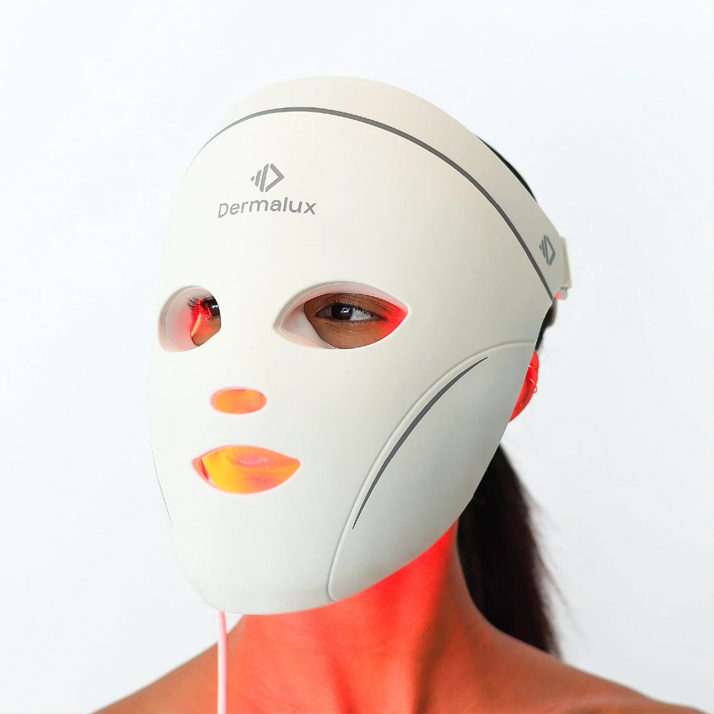 A woman wearing the Dermalux LED Face Mask facing the camera, red light visible through the eye and mouth openings and spilling onto her neck
