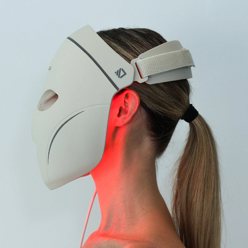 Side view of a woman wearing the Dermalux LED Face Mask, with the adjustable head strap fastened and red light visible along her jaw and neck